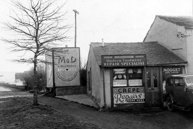 G. Wiggins, boot repairs on Grove Lane. A car is parked on the right. Window advertisements for Nugget Boot Polish and a sign saying, 'Crepe Repairs Done Here' are on the shop.