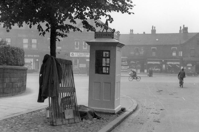 A telephone kiosk on the corner of Spencer Place and Roundhay Road. There is a parade of shops in the background.