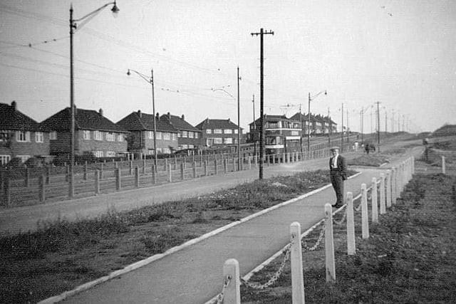 A view looking south along Belle Isle Road towards the junction with Town Street, in the distance on the right. Tramlines run through the centre of the road, where a tram is seen travelling towards Leeds.