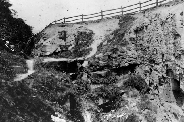 A view of part of old quarry at Belle Isle, shows quarry face with wooden fencing along the top. A man and a boy are seated beside a footpath running beside the quarry.