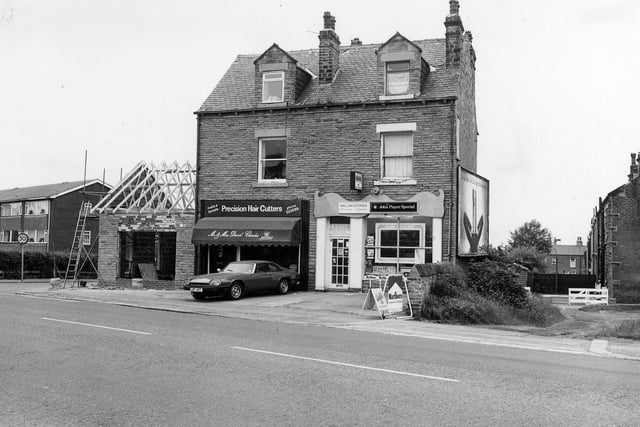 A view of the south side of Bradford Road showing a pair of shops in the centre; to the left is Mr. & Mrs. David Charles, precision hair cutters, and to the right Millan Stores, off licence and cigarettes.