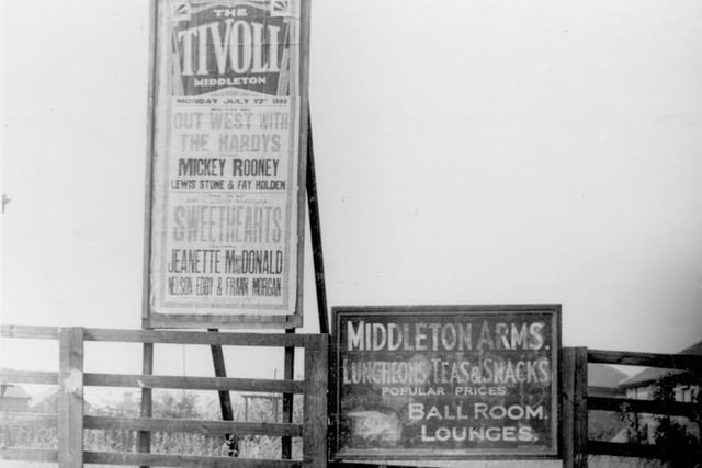 A low timber fence on Middleton Park Avenue. There is a painted wooden sign offering directions to the Middleton Arms, and beside this a free-standing hoarding promoting films at the Tivoli.