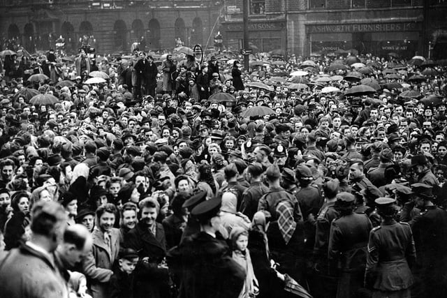 Dense crowds on The Headrow and Victoria Square, in front of Leeds Town Hall. They are waiting for the Victory Parade to begin, part of the VE celebrations. Although a day of heavy rain, there are many smiling faces.