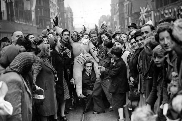 Group of soldiers and civilians celebrating on Briggate. The soldiers are carrying an effigy of Adolf Hitler, a noose is being held up behind the head of the guy. The soldiers are in jovial mood, all carrying bottles of beer, the man on the right is giving a drink to the woman next to him.