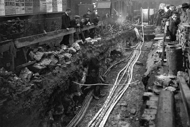 A view along the length of excavations of old cellars in Aire Street, showing piping being laid, workman standing at edge of trench and workman's hut. Also in view is the premises of Rhodes and North.