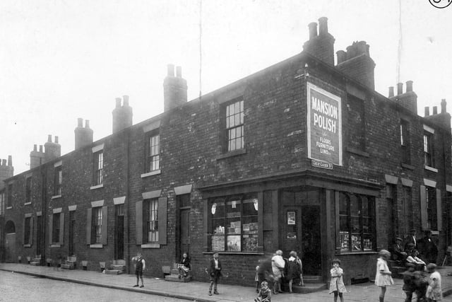 The corner of Great Garden Street and Accommodation Road in Burmantofts, showing corner shop with wall advertising for Mansion Polish; children playing in street and group of men on doorstep.