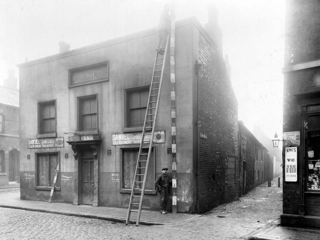 Boarded up 18 Bridge Road, later demolished. Ladder is leaned up against the building and a man is measuring the height of the building for the camera. To the left is an alleyway leading down to Front Walk.