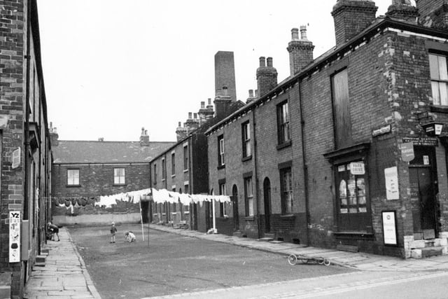 Barker Square from the junction with Commercial Street in Morley. Children are playing on the unmade road and washing hangs between the houses. Cornershops can be seen on either side of the Square.