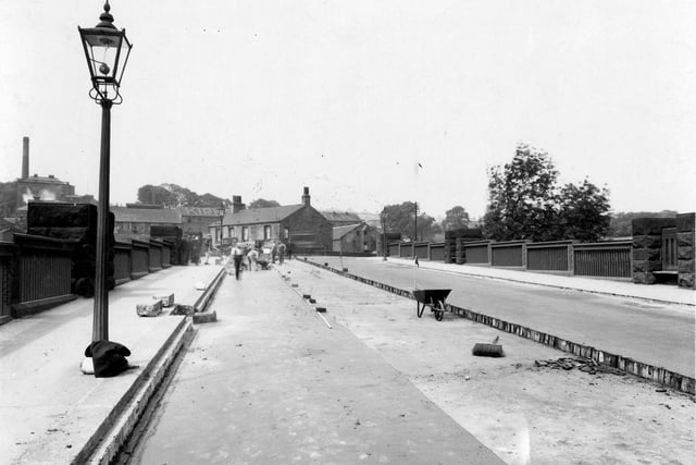Resurfacing of Kirkstall Bridge in June 1938. Men working on the road, wheelbarrow & broom on road. Lamppost on left hand side with some clothing piled up at its base. Kirkstall Brewery & tall chimney in background.