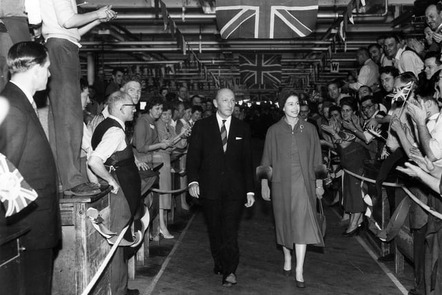HM Queen Elizabeth II during a visit to the factory. She is walking alongside company chairman Lionel Jacobson while workers applaud her on both sides with Union Jacks unfurled above