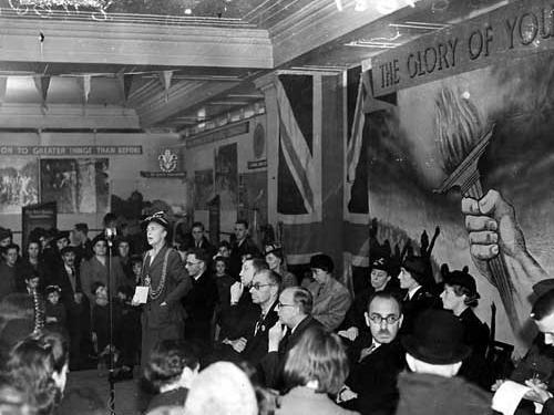 Jessie Kitson opens the Youth Week exhibition in Lewis's store in 1943. The walls are decorated with the slogans and emblems of various youth groups, some of the seated guests are wearing the uniforms of various organisations.