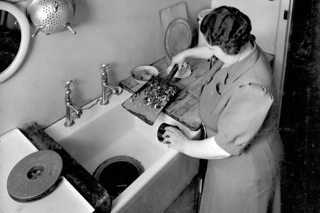 1943 and a lady in the kitchen demonstrates the use of the Garchey waste disposal unit in the sink. Large cover on left has been removed from disposal shaft, she is tipping down a shovel of ashes from a coal fire.