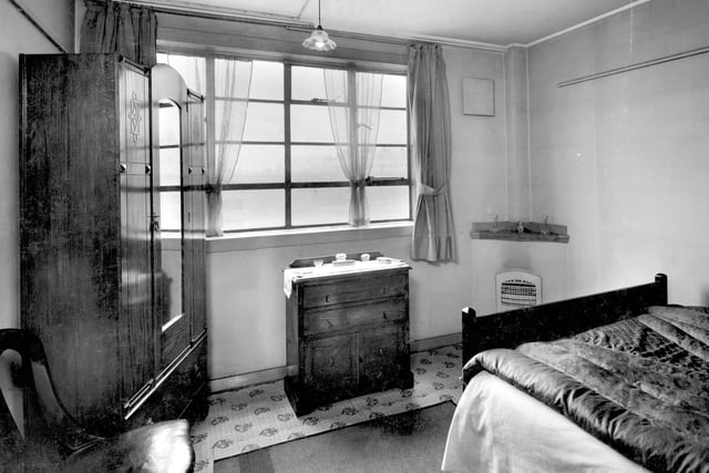 1939. Bedroom furnished with wardrobe, chest of drawers and bed. The bed is covered with a satin eiderdown for warmth. An electric fire is fitted into the corner.
