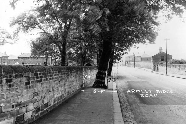 Armley Ridge Road in July 1950. There are stone walls running down both sides of the road. Behind the wall on the right there is a small brick house. In the background there is a row of shops.