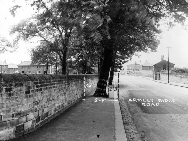 Armley Ridge Road in July 1950. There are stone walls running down both sides of the road. Behind the wall on the right there is a small brick house. In the background there is a row of shops.