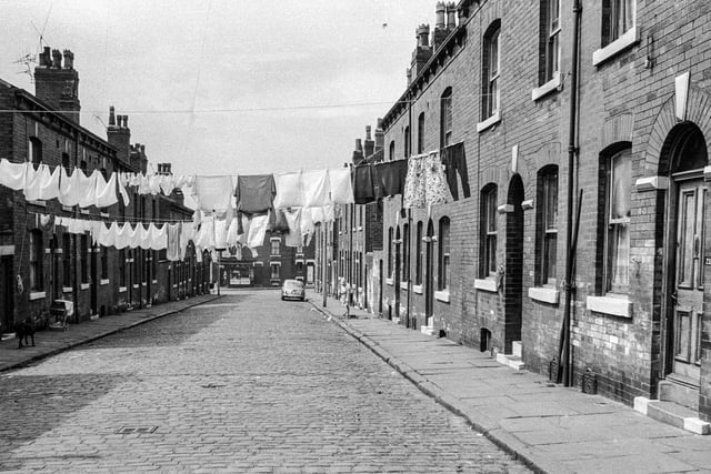 An unmade Leeds street in 1969 with red brick terraced homes either side and lines of washing strung between. The houses have arched entrances with doors opening directly onto the pavement. The gaps between the blocks of properties are likely to house yards with outside toilets.