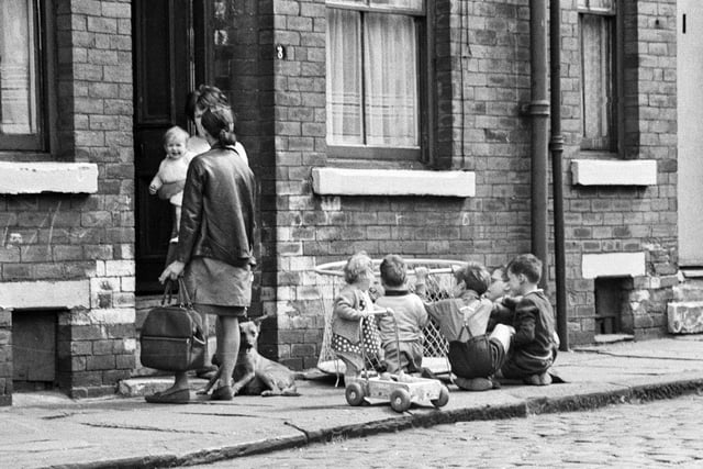 1969. Close up of part of an unknown Leeds street of terraced housing. Two women stand chatting, one is holding a small child while the other carries a shopping bag. Several other children are gathered around a playpen beneath the window