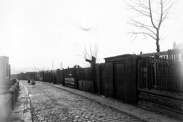 November 1950. View looking south west along Back Cross Green Crescent with two children playing on the cobbled street. The houses have wooden fences and gates. Industrial chimneys can be seen in the background and a drain grate is visible.