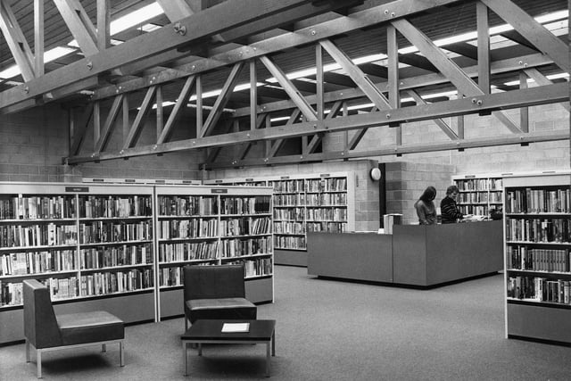 Two members of staff are working at the modern counter and the non-fiction book stock is arranged around the walls. Adult fiction is shelved in free standing bookcases. In the foreground there is comfortable seating and a low table for library users.