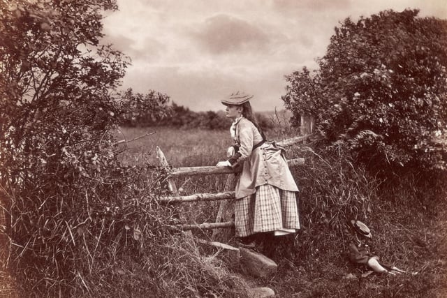 circa 1880: A girl waits next to a stile, as a young child lies asleep in the grass next to her.