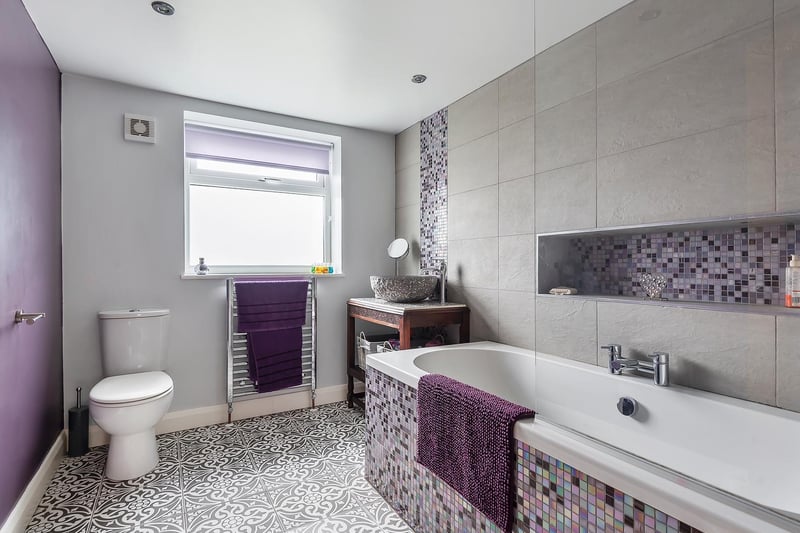 A modern three-piece bathroom suite comprising of a bath with overhead shower, inset shelving, vanity wash hand basin, low level w.c., a frosted double-glazed window, heated towel rail, spot lighting, part tiled walls with mosaic detail and a modern tiled floor.