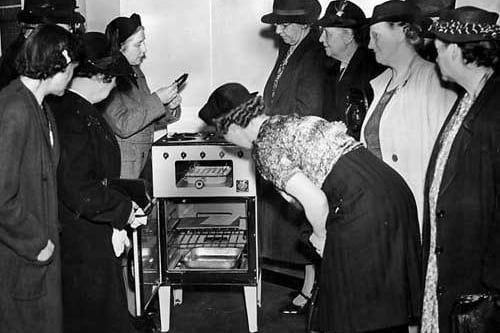 A group of women examine a gas cooker which had four top burners, grill with pan and an oven. It was not unusual for people still to have coal fired ovens and open fires to cook on at this time.