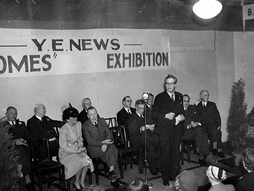 The seated of guests includes a smartly dressed woman in typical 1940s fashion. Hats were commonly worn, this woman is wearing one with a single feather trim. The men have three piece suits which include a waistcoat.