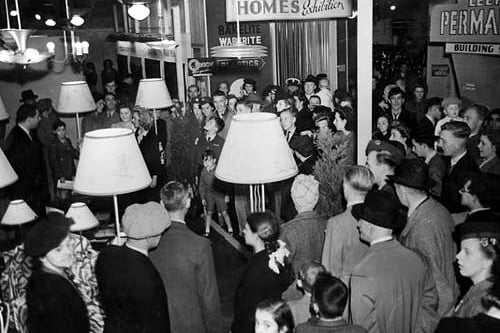 The crowd admire a display of light fittings and lamps. One lady is wearing a scarf wound onto her head in 'turban' fashion. This was widely adopted during the war to contain and protect the hair of women working in the factories.