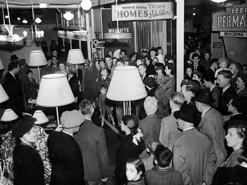 The crowd admire a display of light fittings and lamps. One lady is wearing a scarf wound onto her head in 'turban' fashion. This was widely adopted during the war to contain and protect the hair of women working in the factories.