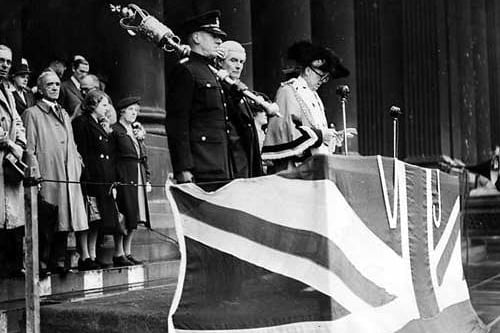 The Lord Mayor of Leeds, Charles Walker stands at a podium draped with the Union Jack flag. He is reading the announcement of the Japanese surrender, bringing an end to the hostilities.