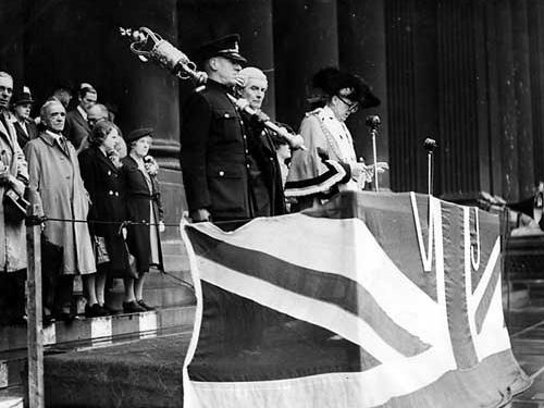 The Lord Mayor of Leeds, Charles Walker stands at a podium draped with the Union Jack flag. He is reading the announcement of the Japanese surrender, bringing an end to the hostilities.