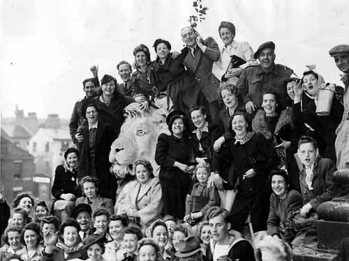 A group of people celebrating outside Leeds Town Hall. One of the lions flanking the entrance is in the middle of the group. Although it was summer time it rained quite heavily, the ladies are wearing scarves or hats.