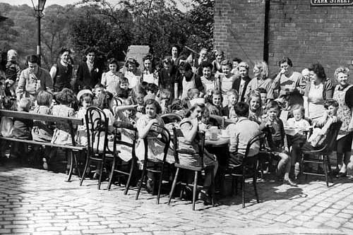 A street party on Park Street in Leeds to celebrate VJ Day. The children are seated round the tables on a variety of chairs and benches, with the women serving their food.