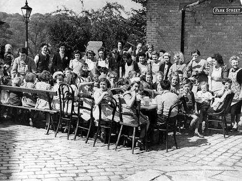 A street party on Park Street in Leeds to celebrate VJ Day. The children are seated round the tables on a variety of chairs and benches, with the women serving their food.