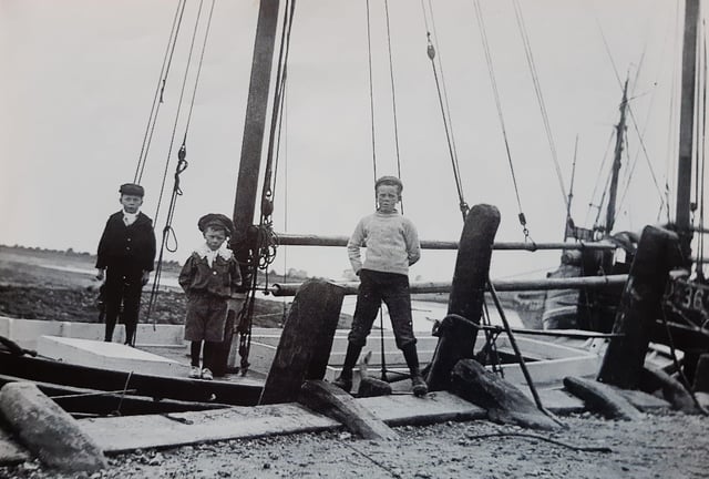 Three smart young men on a boat at Bosham Quay in about 1909. It poses a mystery, as the tide has gone out yet the boat is near the top of the quay. Was the boat on the deck of a much larger craft?