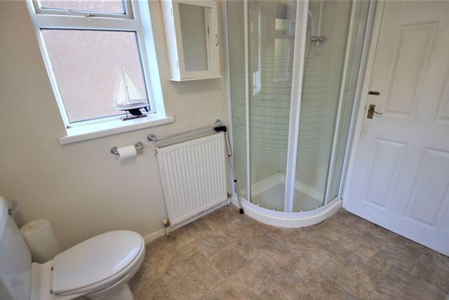 A second view of the bathroom which shows the enclosed shower cubicle tucked away in the corner. The room also has cushioned vinyl flooring, part-tiled walls, a radiator, dual ceiling-lights, and uPVC windows to the side and back.