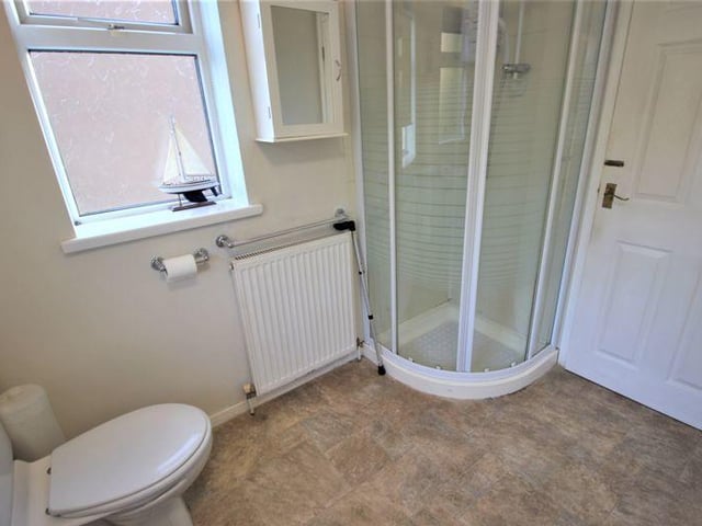 A second view of the bathroom which shows the enclosed shower cubicle tucked away in the corner. The room also has cushioned vinyl flooring, part-tiled walls, a radiator, dual ceiling-lights, and uPVC windows to the side and back.