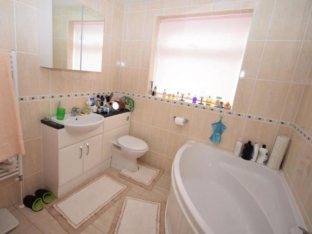 This is the en suite bathroom to the main bedroom. It is fitted with a panelled bath that has a shower above, a low-flush WC and wash basin in a vanity unit that also offers storage. The room is finished with tiled walls and floor.