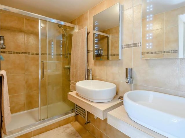 This is the en suite shower room to the master bedroom. A contemporary four-piece suite with chrome fittings includes a tiled shower enclosure, his and hers wash hand basins, fitted mirrors above each sink with inset lighting, a low-flush WC and a towel rail.
