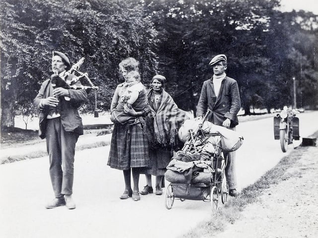 A piper escorts a travelling family, with some of their possessions - including a set of pipes - wheeled along in the pram.