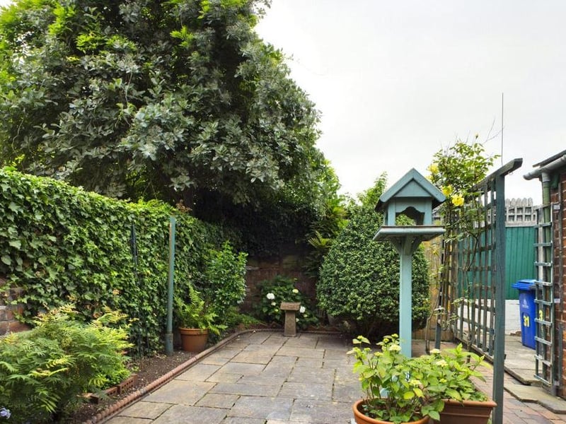 Rear garden - Wall enclosed with timber access gate opening onto the rear service path; paved with raised flower beds and a variety of mature plants, flowers and shrubs, tool store and garden taps.