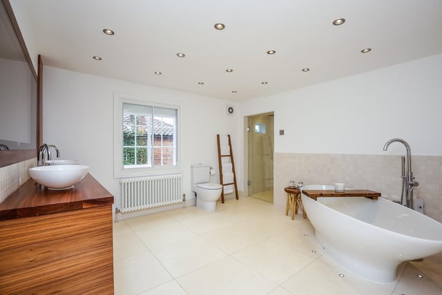 Double ended bath with surrounding uplighting, walk in rainfall shower, close coupled WC and twin wash hand basins on cabinet with drawers beneath. Wall mounted TV concealed within a mirror above, tiled floor with underfloor heating.
