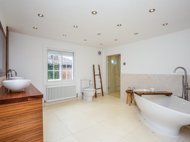 Double ended bath with surrounding uplighting, walk in rainfall shower, close coupled WC and twin wash hand basins on cabinet with drawers beneath. Wall mounted TV concealed within a mirror above, tiled floor with underfloor heating.