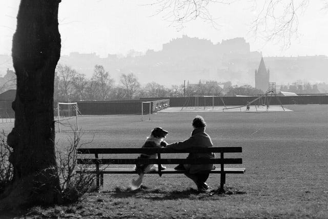 A woman and her dog sit on a bench in Inverleith Park enjoying the view of Edinburgh Castle in March 1961.