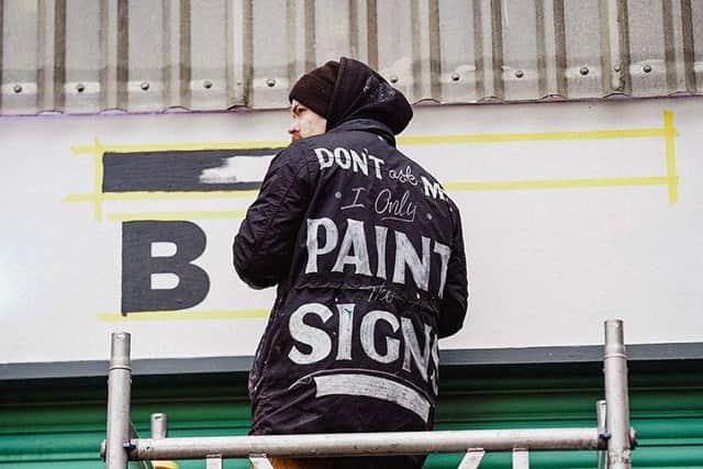 Tatch Hatch-Robertson at work wearing his a jacket that says 'don't ask me, I only paint the signs' written on the back. Picture: Tatch Hatch-Robertson