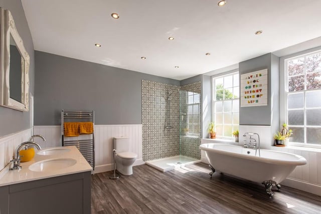 This bathroom features a freestanding roll top bath, a step-in shower with a fixed glass screen and twin wash hand basins with vanity cupboards beneath. There is a walk-in towel cupboard, a vertical radiator, frosted sash windows and a heated towel rail.