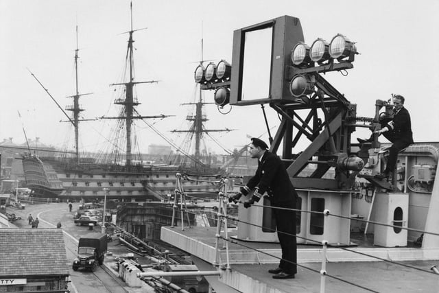 Naval officers check the new mirror deck landing aid for aircraft on HMS Victorious after the warship was extensively reconstructed and modernised in January 1958, while HMS Victory sits in her dry dock as a museum ship.