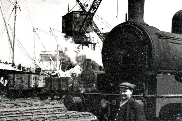 This photos shows a steam engine and driver on the docks with a crane unloading timber in the background. Photo: Hartlepool Library Service.