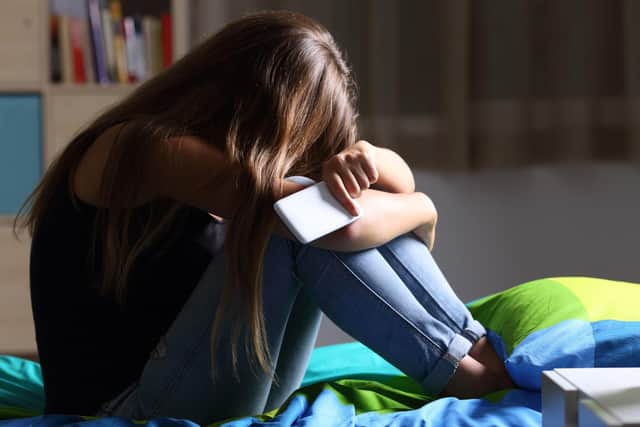 A stock picture of a teenage girl sitting on a bed holding a mobile phone, head in hands.
