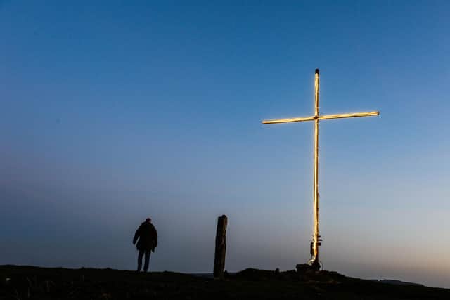 A walker looks on across Stocksbridge from the cross on a hill above the valley as the sun sets.
